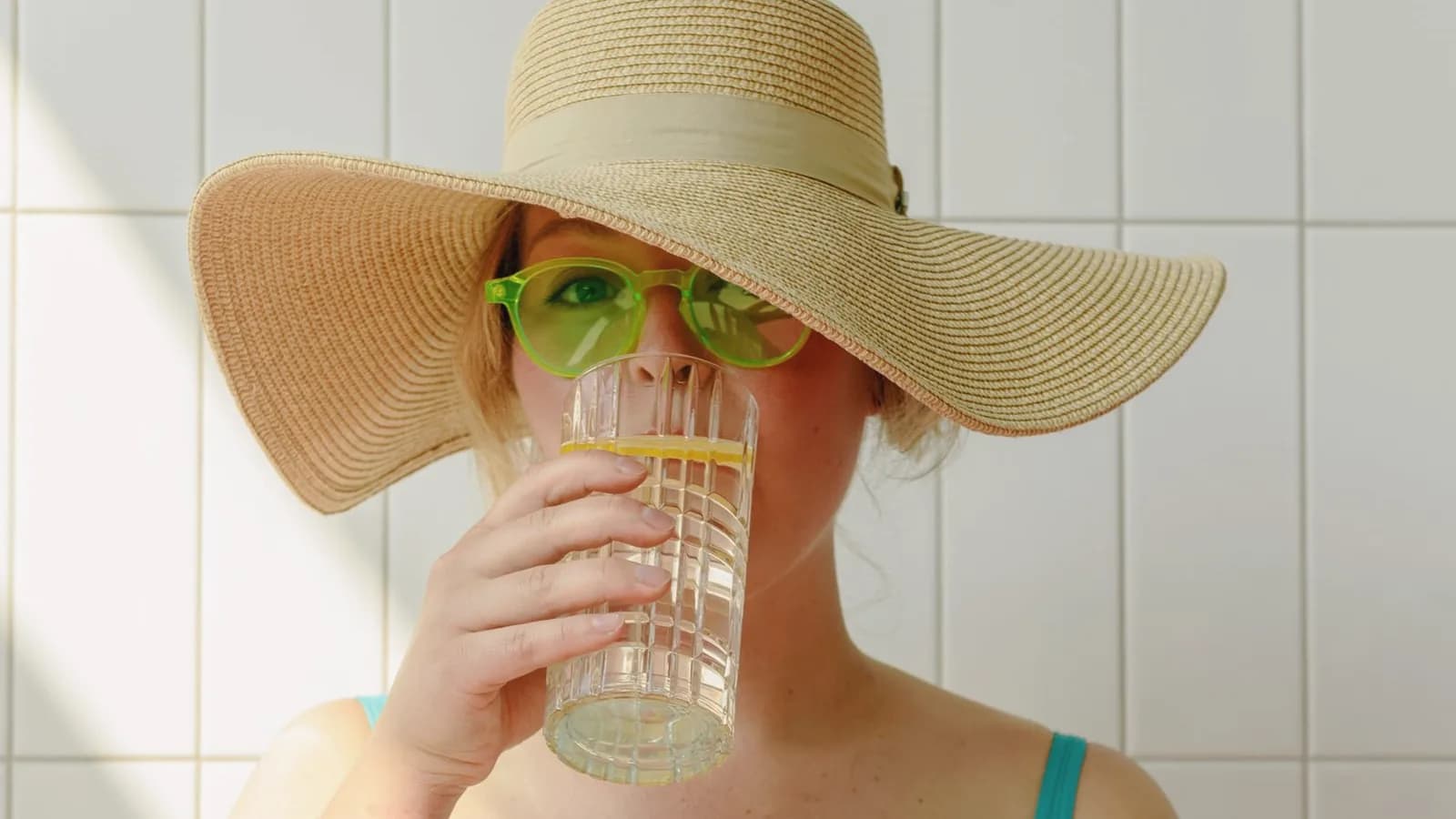 Hydration during pregnancy before an ultrasound: Woman in a straw sun hat drinking a glass of lemon water in a bright tiled