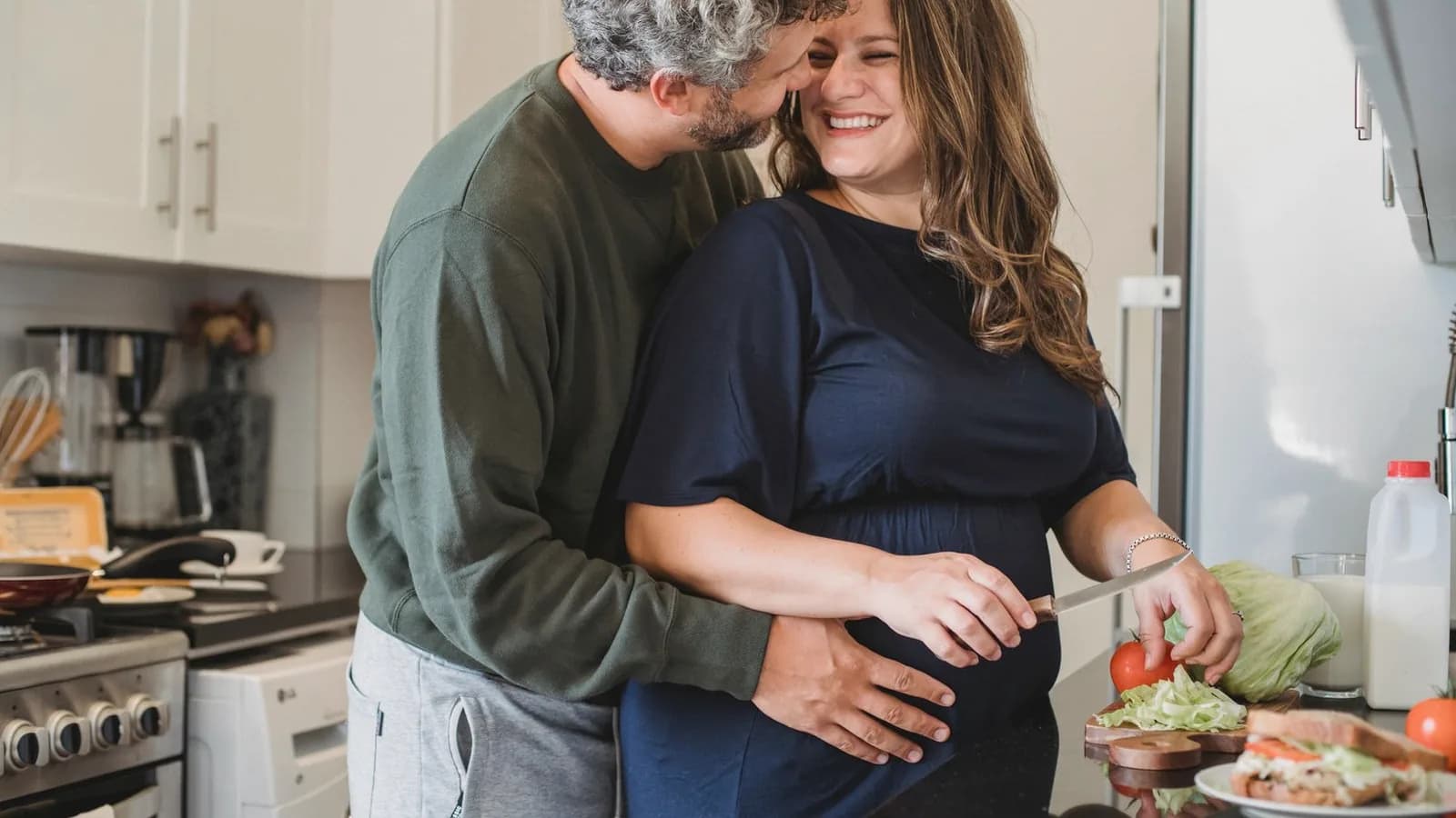 Involve partner in pregnancy: Husband embracing smiling pregnant wife while they prepare food together in a kitchen