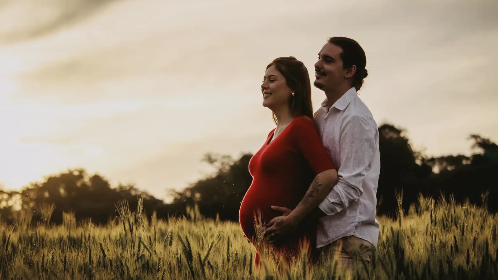 Strengthening relationship during pregnancy: Smiling couple embracing in a golden wheat field at sunset, woman in red dress