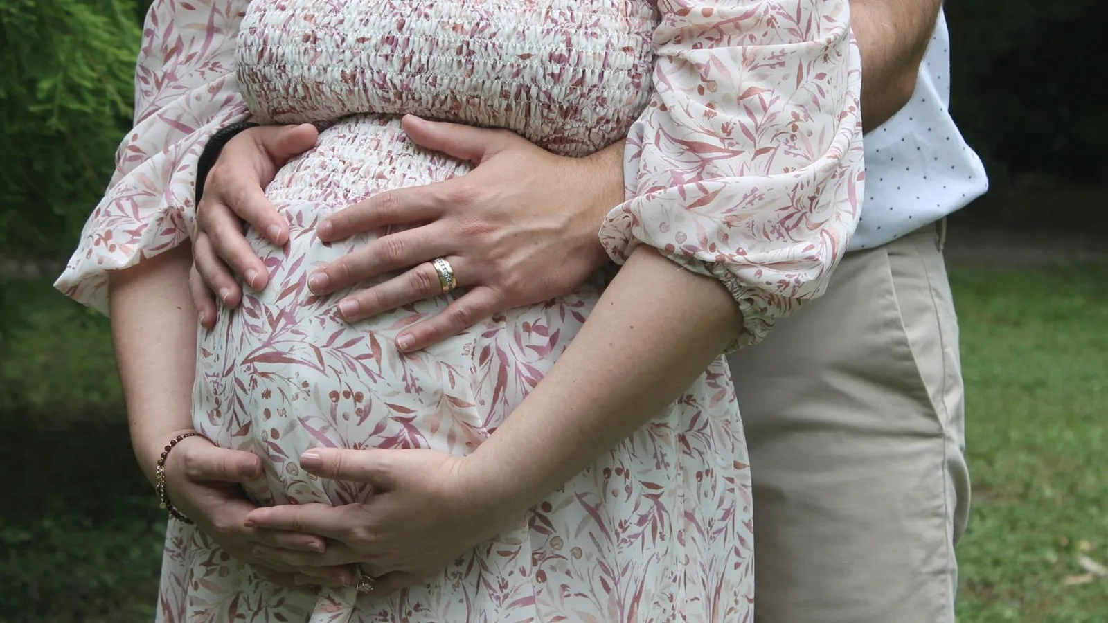 How to support a pregnant partner: Couple gently holding a pregnant belly in a floral dress on green lawn