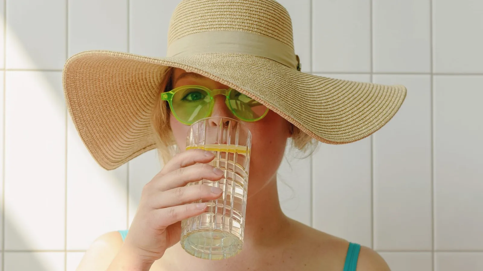 Hydration during pregnancy before an ultrasound: Woman in a straw sun hat drinking a glass of lemon water in a bright tiled