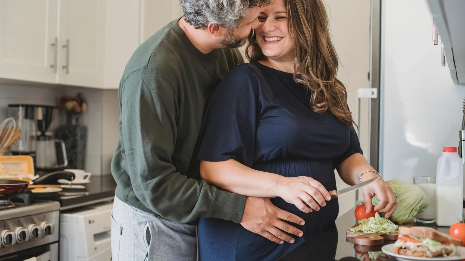Involve partner in pregnancy: Husband embracing smiling pregnant wife while they prepare food together in a kitchen