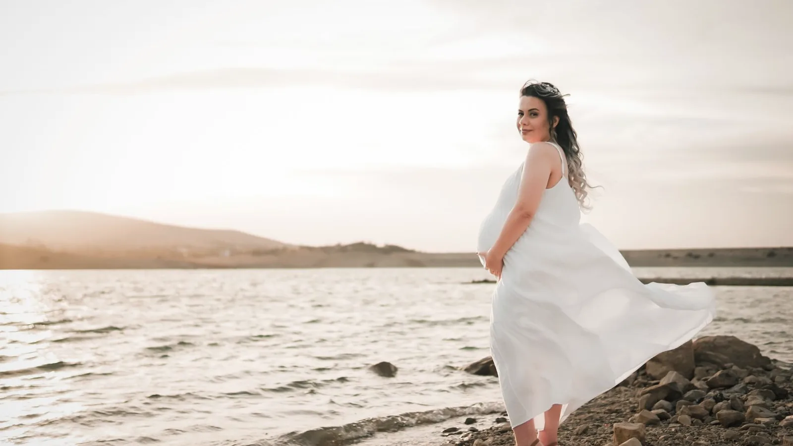 Maternity photoshoot vs ultrasound session: Pregnant woman in a flowing white dress posing by a lake at sunset