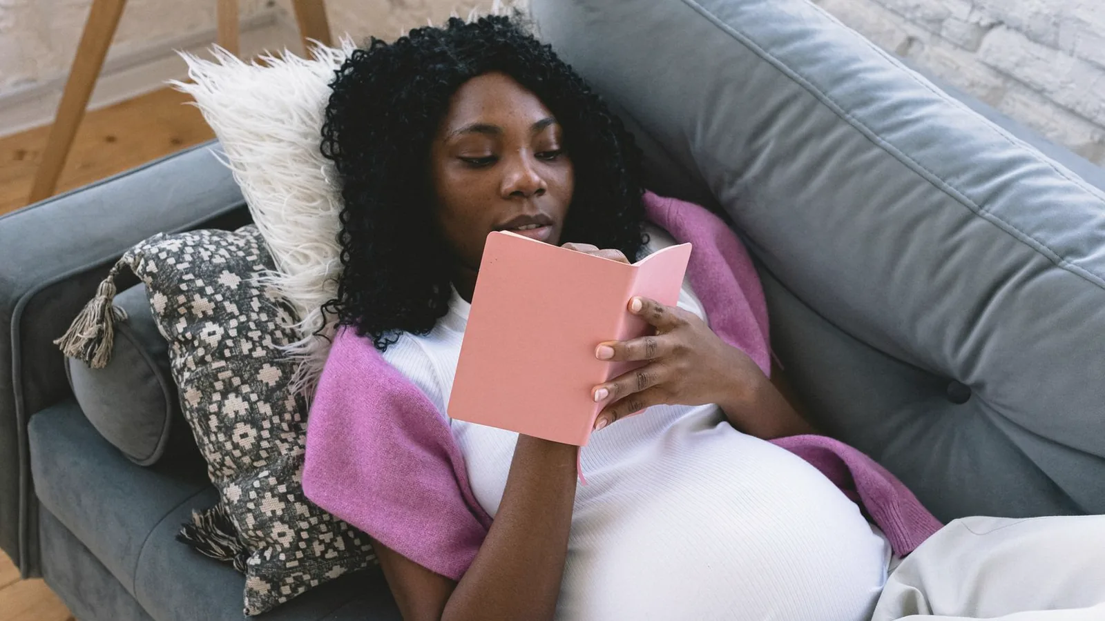 Pregnancy self care ideas: Pregnant woman relaxing on a sofa with a blanket, reading a pink notebook