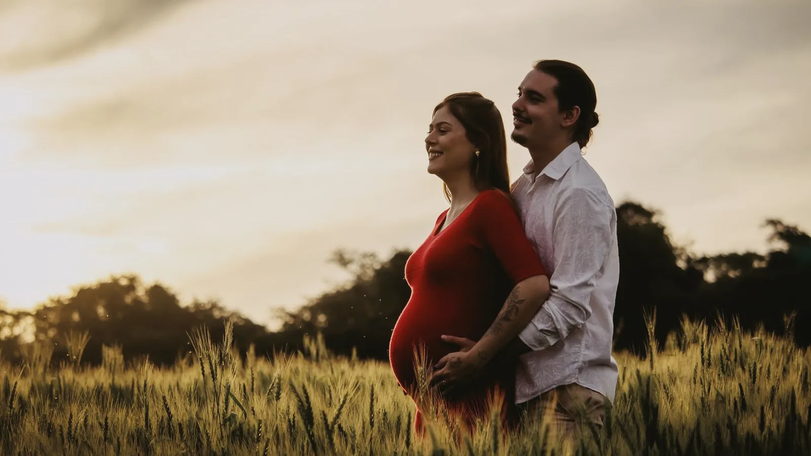 Strengthening relationship during pregnancy: Smiling couple embracing in a golden wheat field at sunset, woman in red dress